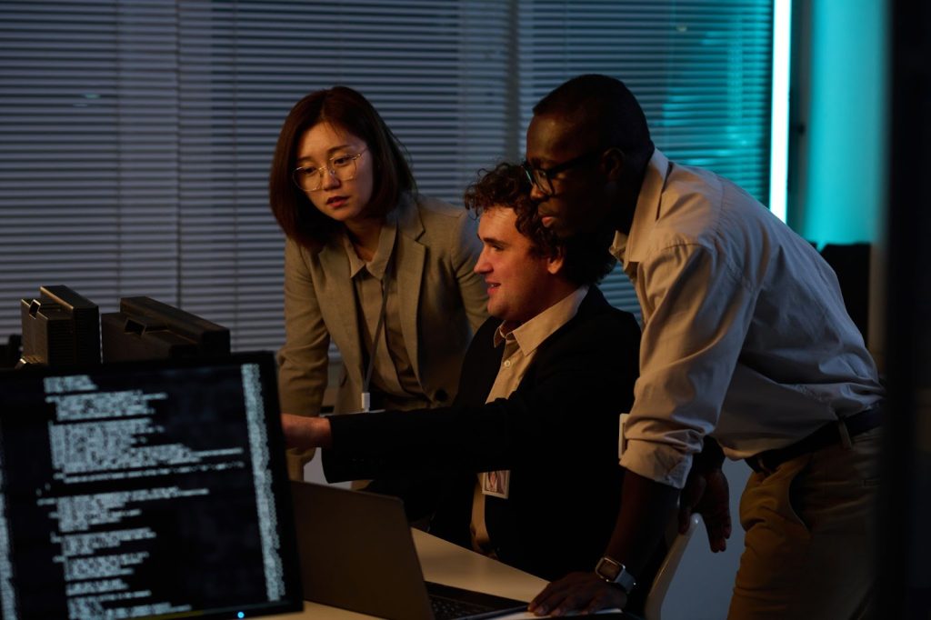 Two men and a woman in business attire stare at a computer screen next to a monitor displaying code.
