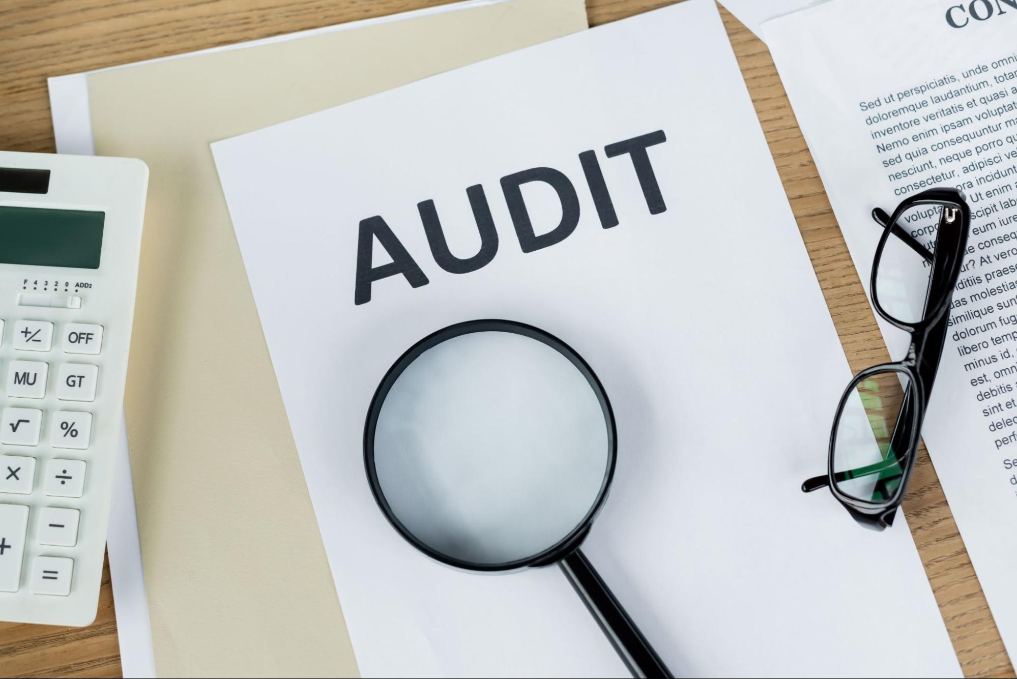 A top view of a magnifying glass resting on a sheet of paper labeled "Audit," positioned next to a calculator and eyeglasses, representing a compliance audit.