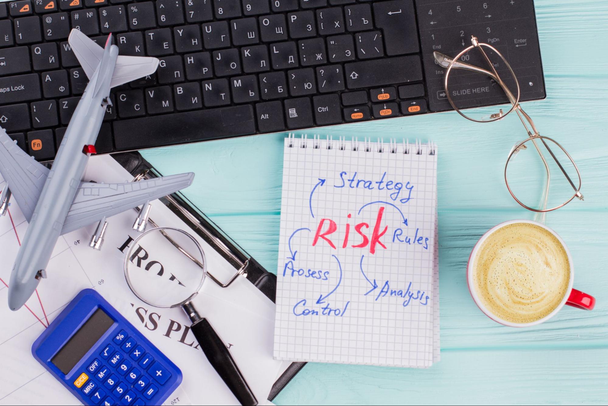 An office desk with a cup of coffee, eyeglasses, computer keyboard, calculator, and toy airplane, alongside a note on risk and strategies, reflecting risk mitigation planning.