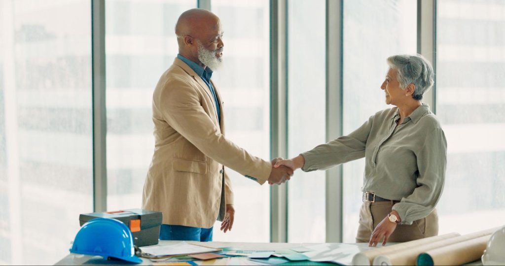 A pair of mature business professionals shake hands in a high-rise, glass-walled office, sealing a successful partnership following a productive B2B collaboration.

