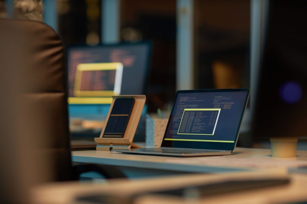 A smart phone and a laptop with coding on their respective screens sitting on a desk in front of a chair.
