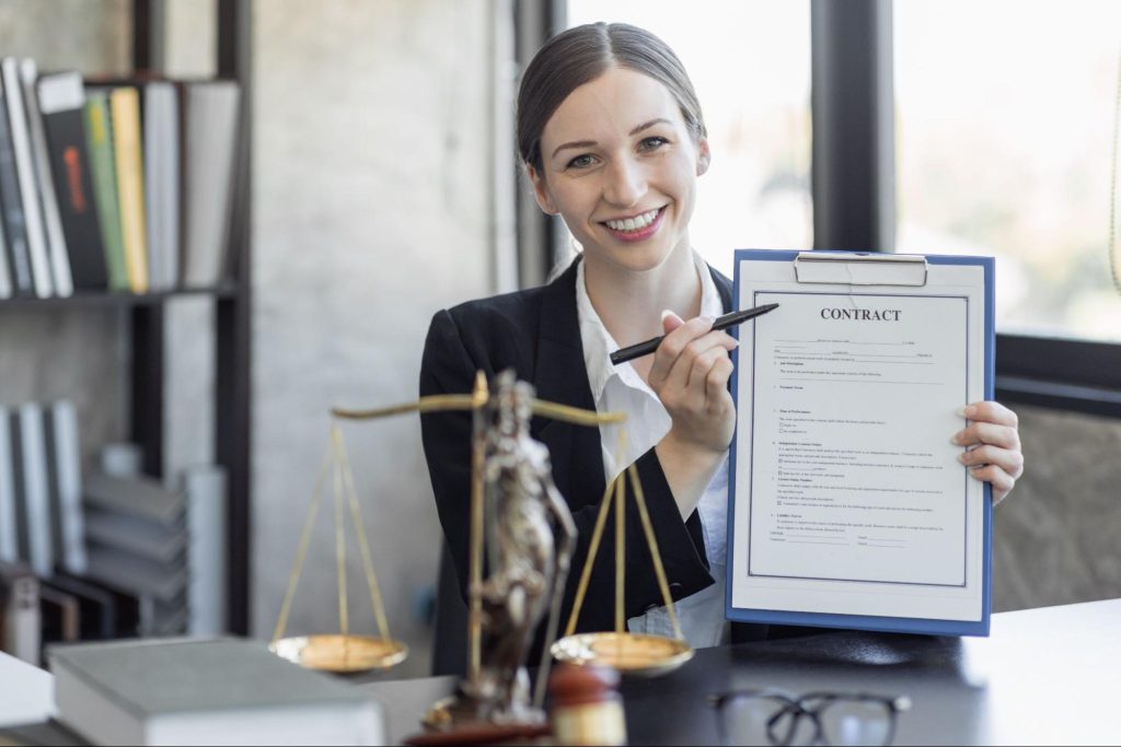 A female lawyer in a suit is at her workplace, pointing at a legal document she’s holding. On her desk are a statue of Lady Justice, a laptop, a gavel, and scales of justice. Behind her is a large glass window and a bookshelf filled with books.

