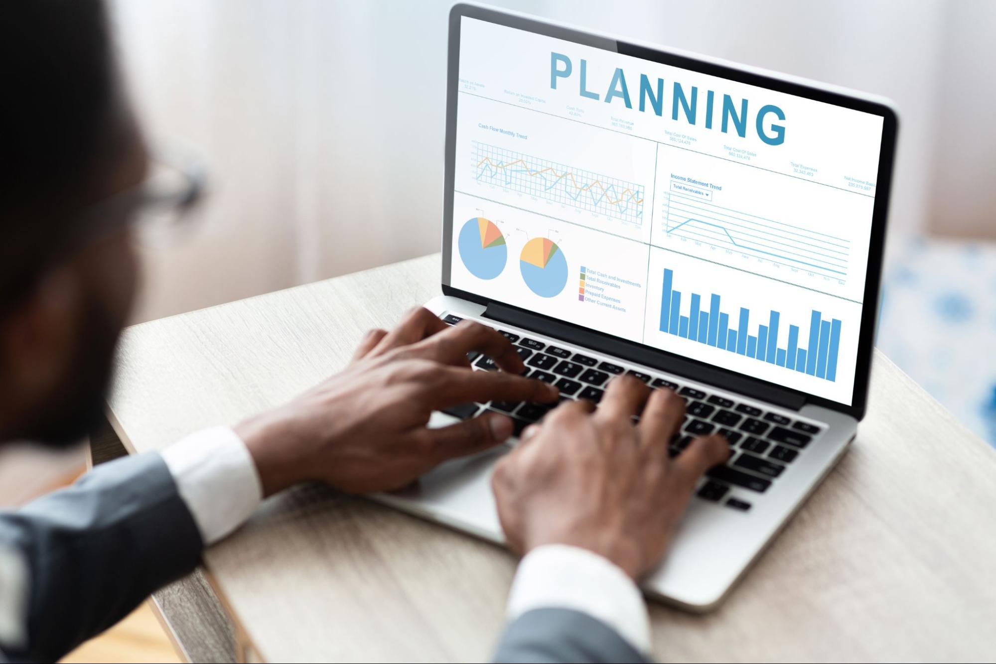 An African businessman working on a laptop, analyzing financial graphs and charts displayed on the screen. The focused setting highlights strategic business planning.