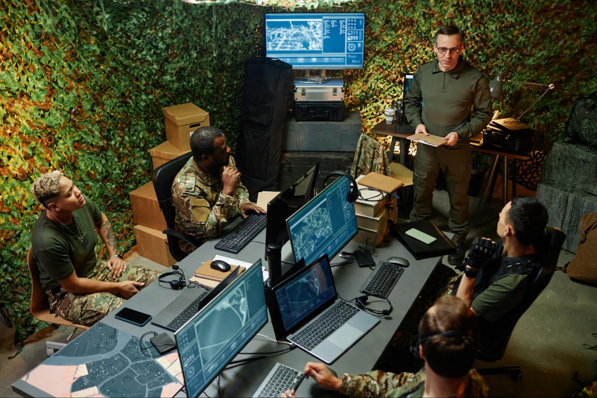 A group of young, intercultural officers in camouflage uniforms attentively listening to their superior's report in a computer room at their headquarters.