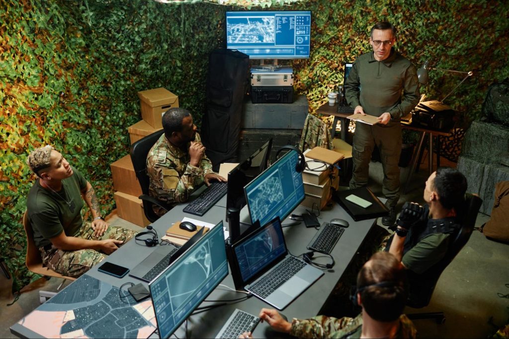 A group of young, intercultural officers in camouflage uniforms attentively listening to their superior's report in a computer room at their headquarters.
