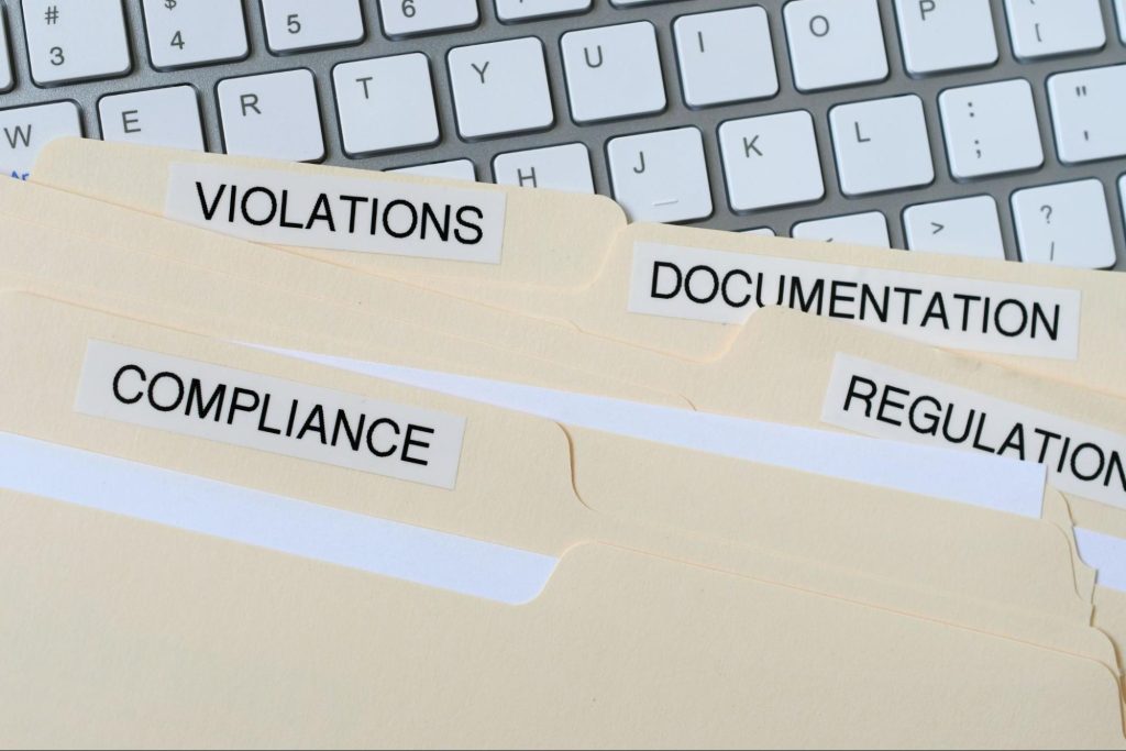 A close-up shot of file folders labeled Compliance, Violations, Documentation, and Regulations placed on a white computer keyboard.