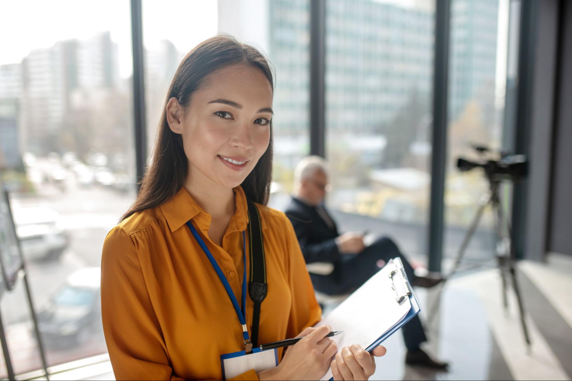 In a business office, a woman with long dark hair is wearing a yellow shirt, writing on a clipboard, and smiling into the camera.