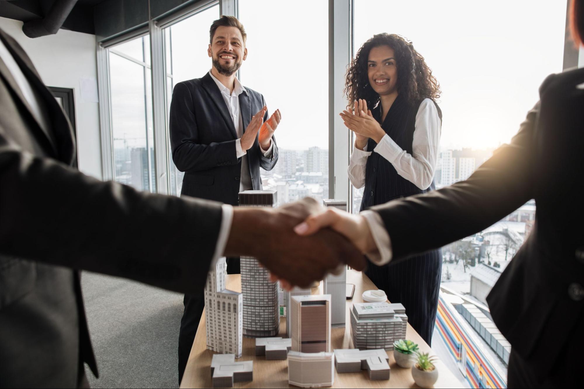 Two business professionals shake hands at an office while other company members clap in the background.