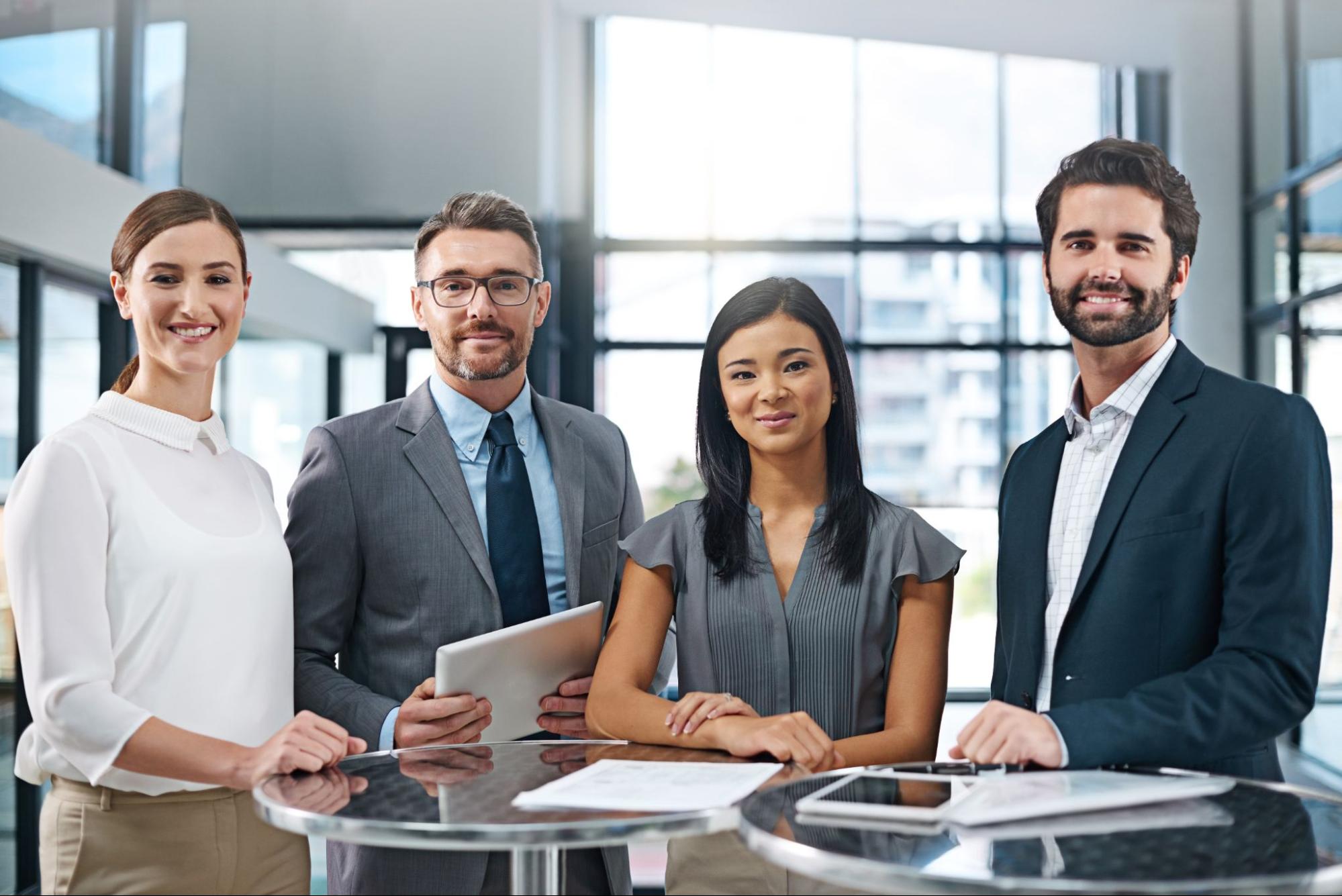 Four businesses people standing next to two glass tables.