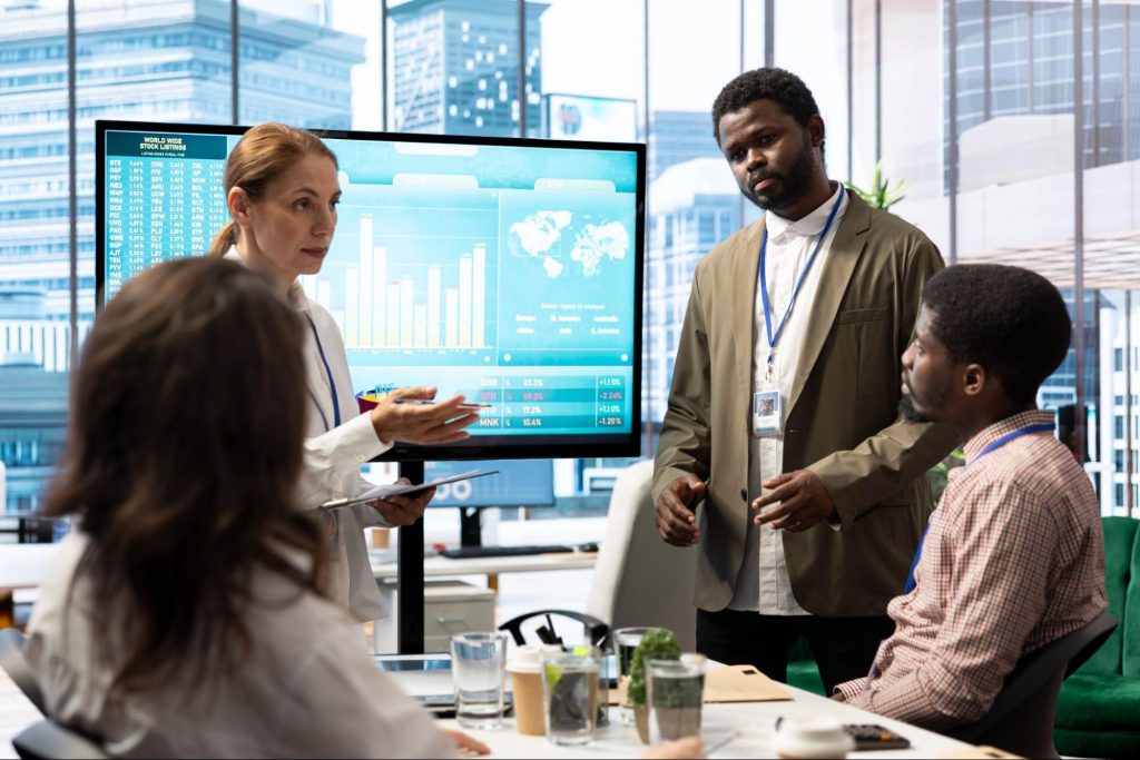 A female financial expert briefs her team on new enterprise objectives, presenting data on a screen in a conference room with glass windows overlooking the city skyline.
