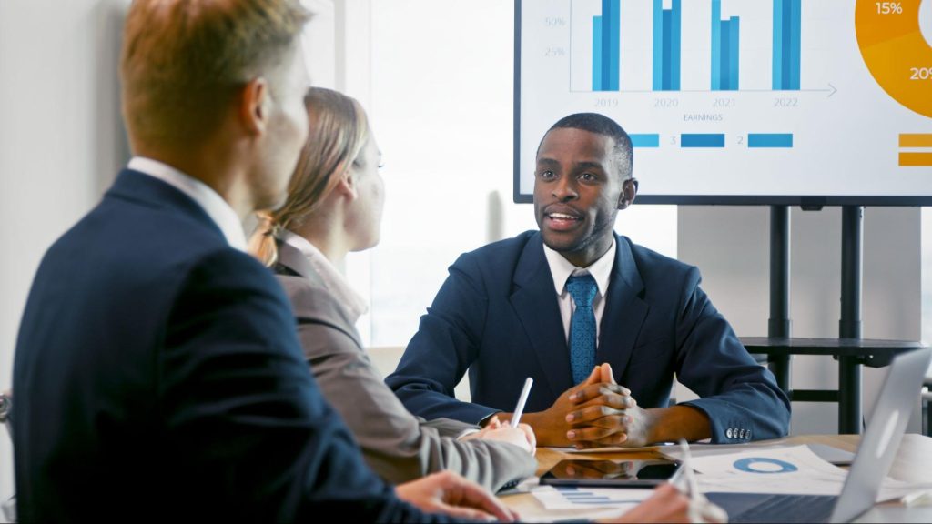 A young Black financial consultant presents a company's profit analysis to clients inside the boardroom.